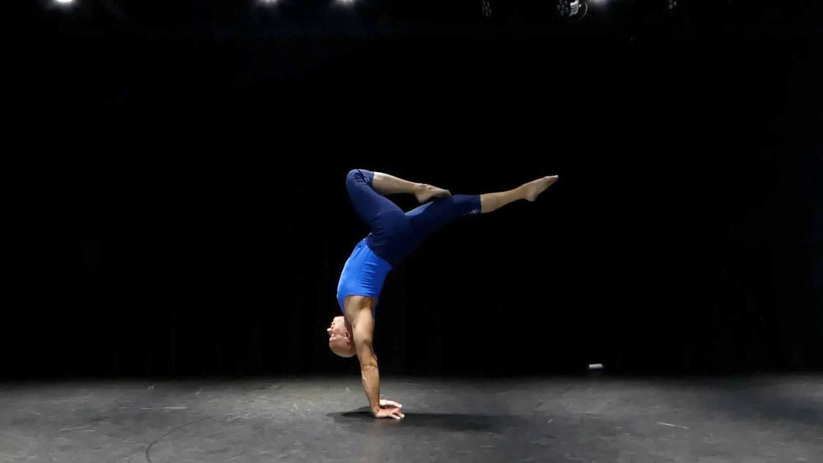 Ben Lowrey alone on a dark stage in a split-leg handstand, one knee bent and the other leg reaching out behind him
