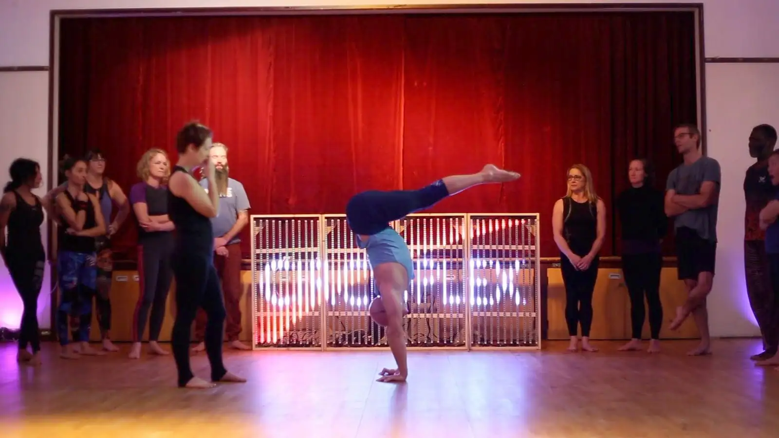 A student holding a handstand in front of the CIRCOPLEX LED sign and red curtain, with a crowd around the edge