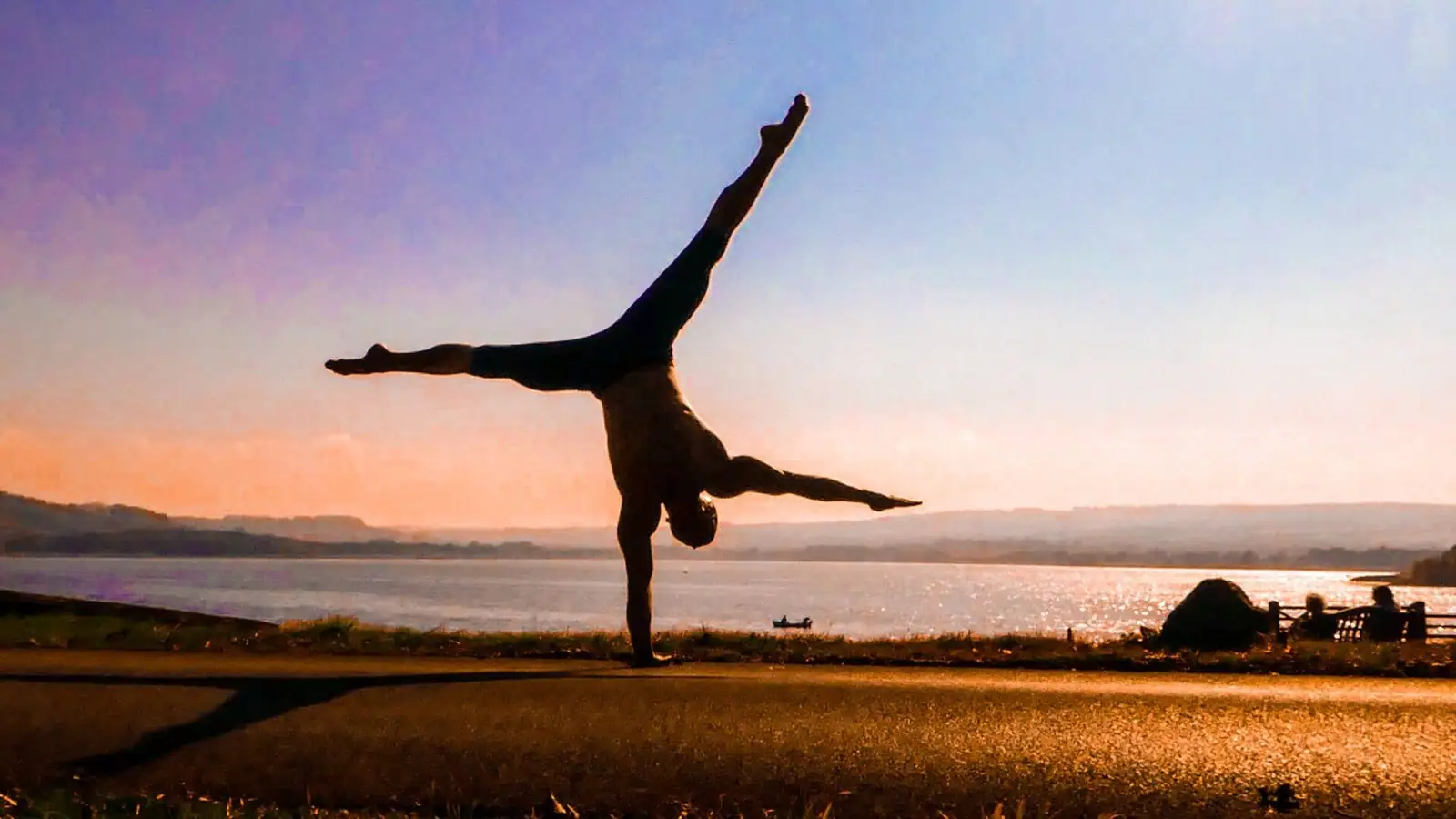 Silhouette of Ben Lowrey in a one-arm handstand against an orange sunset over a lake